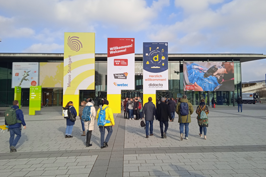 Vorplatz der didacta 2025 in der Messe Stuttgart. Menschen laufen auf den Eingang zu und es gibt verschiedene Schilder u. a. eins mit didacta Logo und Infos zur Messe